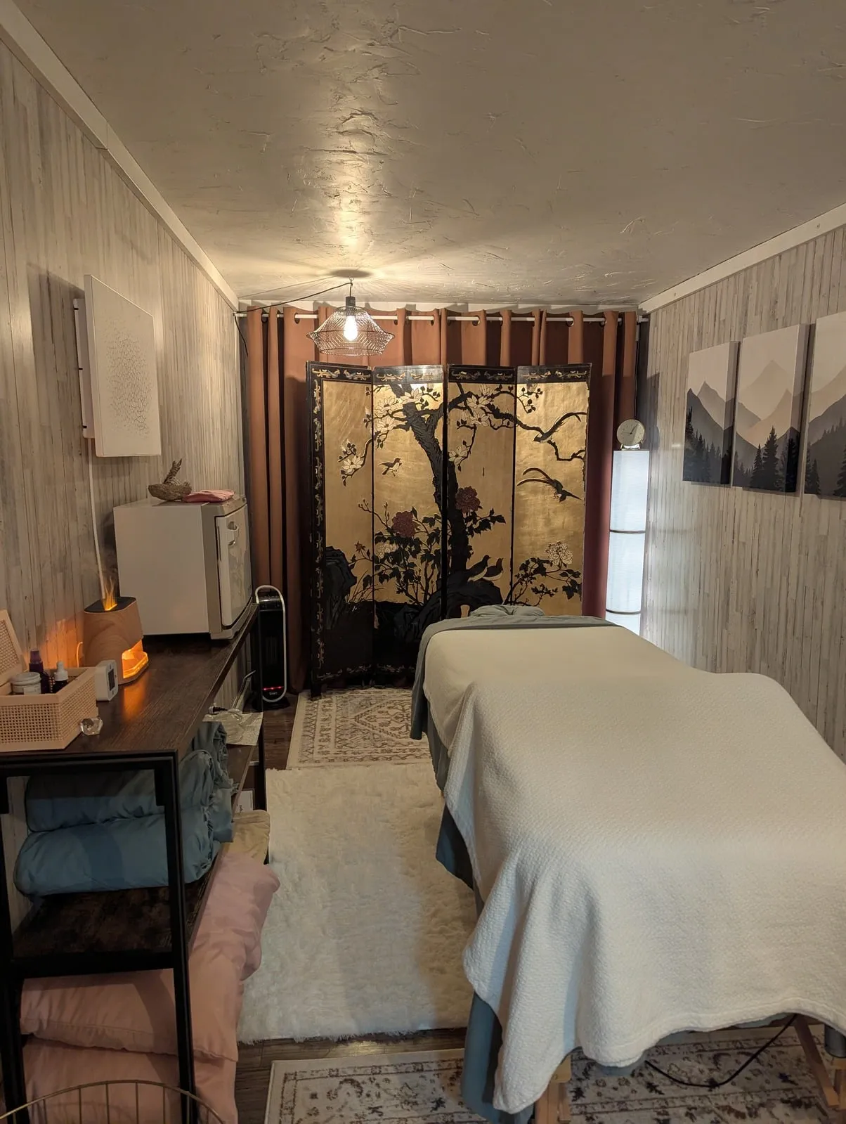 Massage treatment room in Granby with a heated massage table, gold lacquer floral screen, warm pendant lighting, and mountain artwork
